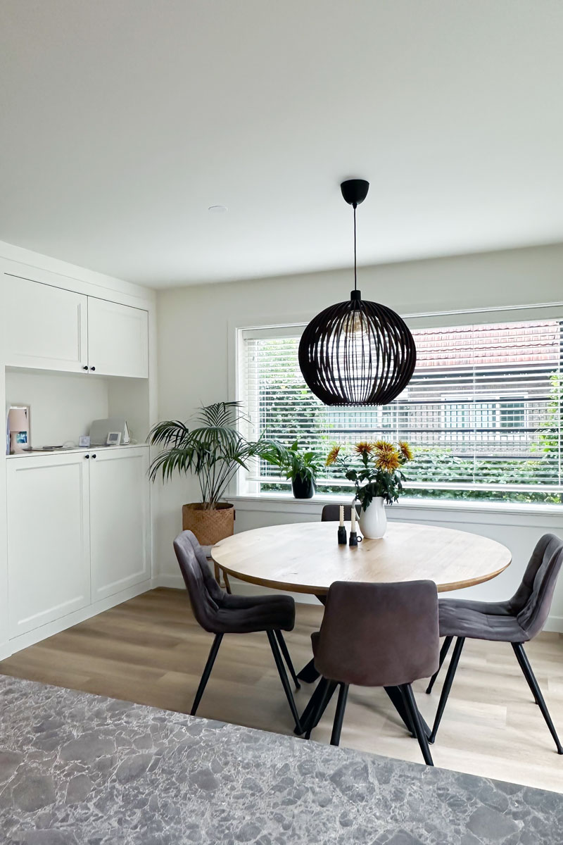 Lichte eetkamer in jaren 30 woning met ronde tafel, groene planten en grote raampartijen.